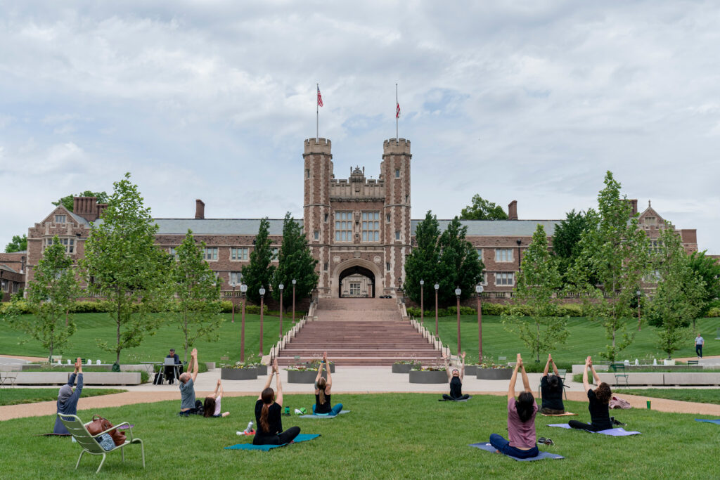 A group of people practice yoga on mats on a grassy lawn in front of a large, historic university building with two towers and flags, under a cloudy sky.