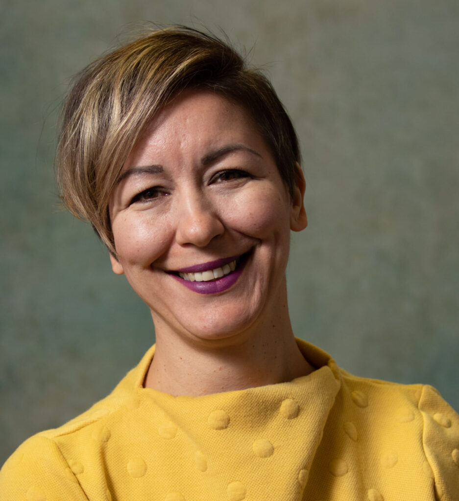 A smiling person with short, light brown hair and purple lipstick wears a yellow textured top, standing in front of a neutral, greenish background.