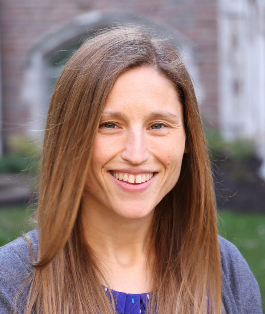 A woman with long, straight light brown hair smiles outdoors. She is wearing a blue top and a gray sweater, with a blurred background of greenery and a brick building.