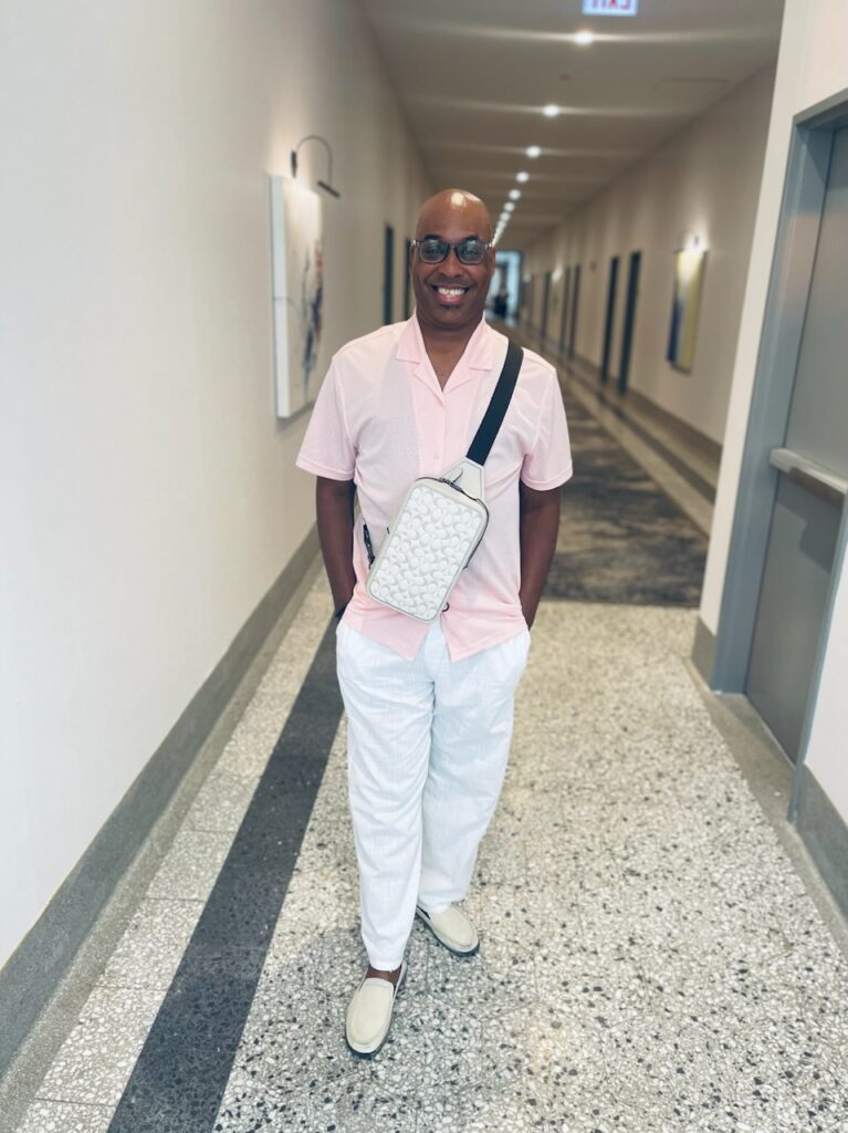 A smiling person in a light pink shirt and white pants stands in a brightly lit hallway, wearing glasses and a patterned crossbody bag. The hallway has artwork on the walls and doors on either side.