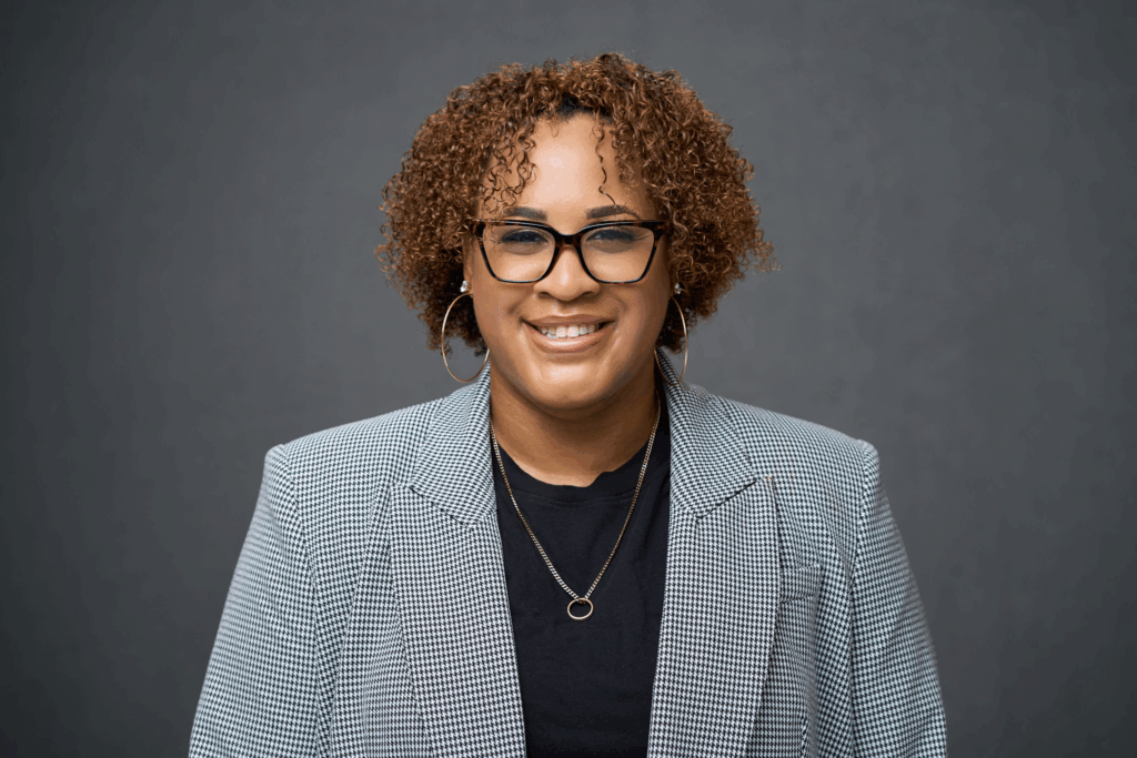 A woman with curly hair, glasses, and hoop earrings smiles at the camera. She is wearing a black top, a checkered blazer, and layered necklaces, standing in front of a plain gray background.