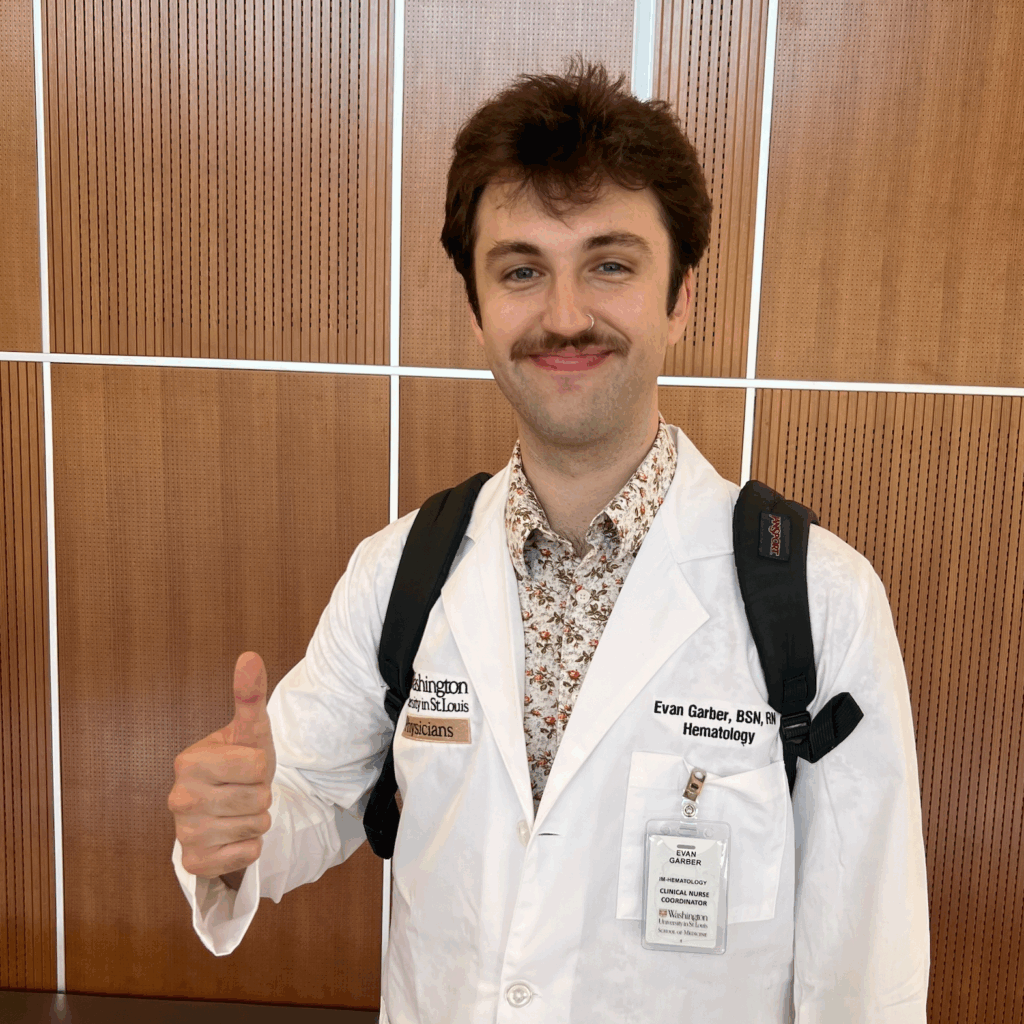 A person with brown hair and a mustache, wearing a white lab coat and floral shirt, smiles and gives a thumbs up. They have a backpack and ID badge and stand in front of a wooden wall with vertical white lines.