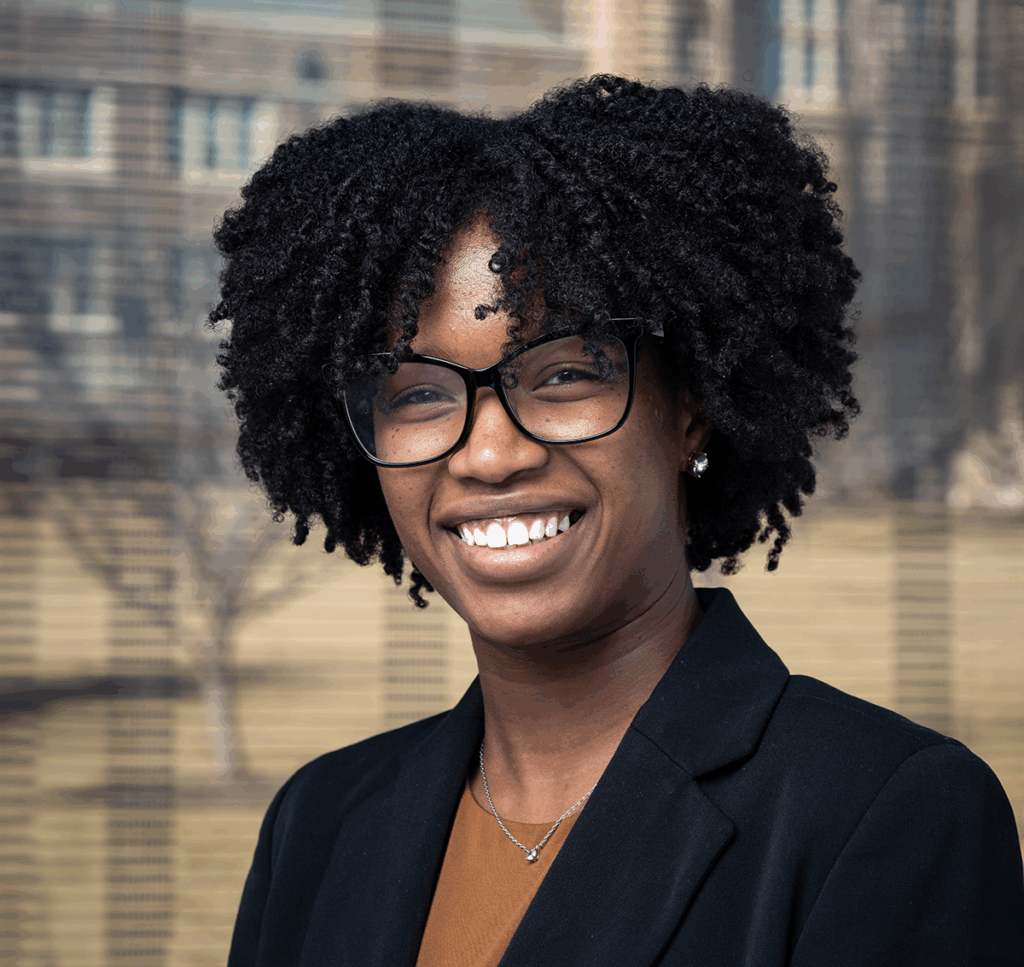 A woman with curly black hair and glasses, wearing a black blazer and a brown top, smiles while standing outdoors with blurred buildings and trees in the background.