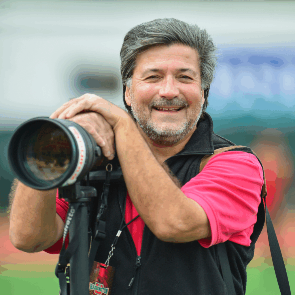 A smiling man with gray hair and a beard leans on a large camera with a telephoto lens mounted on a tripod. He is wearing a red shirt and black vest, and is outdoors with a blurred background.