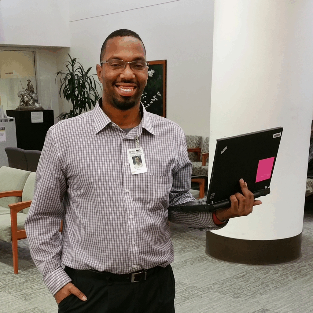 A man wearing glasses, a checked shirt, and an ID badge stands indoors, smiling and holding a closed laptop with a pink sticky note on it. The background includes chairs, plants, and office decor.