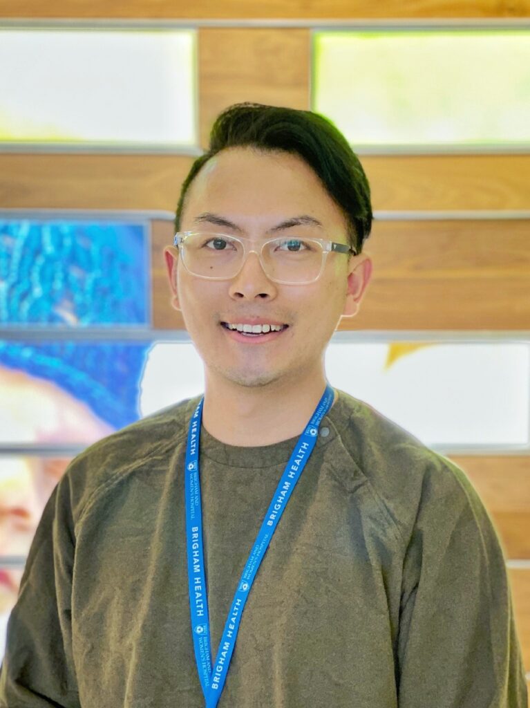 A person with short, dark hair and glasses smiles at the camera, wearing a brown shirt and a blue lanyard that reads Bingham Health. The background features wooden panels and colorful artwork.
