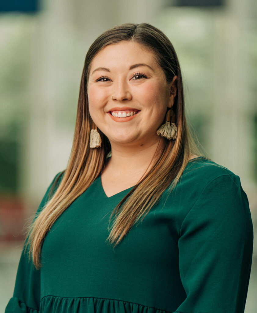 A woman with straight, light brown hair wearing a dark green blouse and tassel earrings smiles at the camera in a softly lit indoor setting.