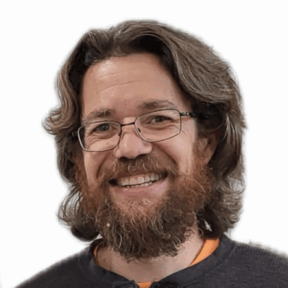 A smiling man with long wavy brown hair, a full beard, glasses, and wearing a dark sweater, posing against a plain white background.