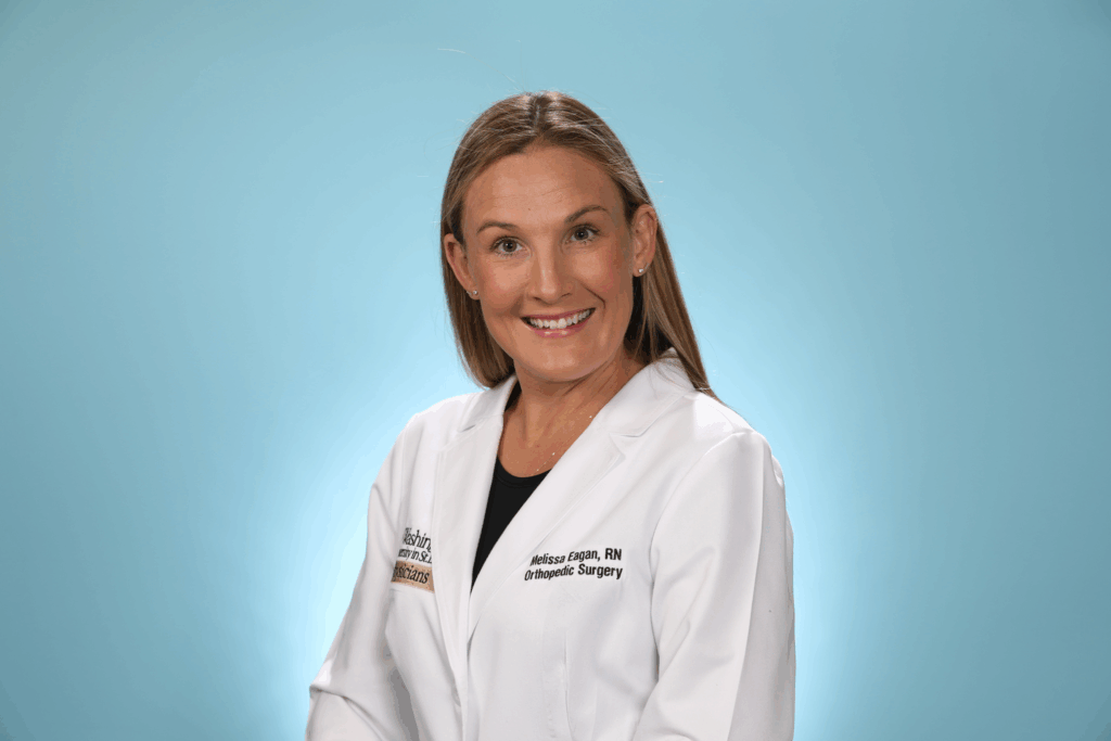 A woman with straight blonde hair, wearing a white medical lab coat, smiles in front of a plain blue background. The coat has embroidered text and a badge on it.