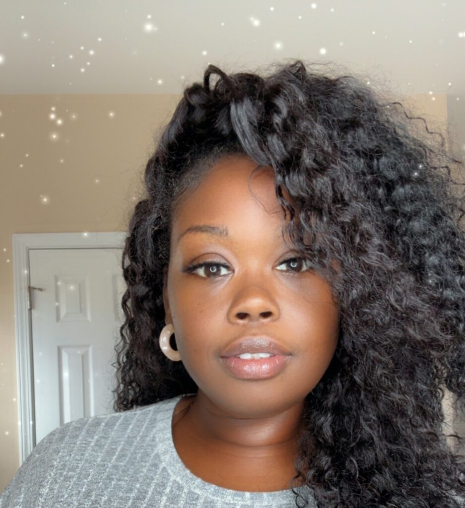 A woman with curly hair wearing hoop earrings and a gray textured top looks at the camera. The background is a beige room with a white door, and soft, glowing sparkles are overlaid on the image.