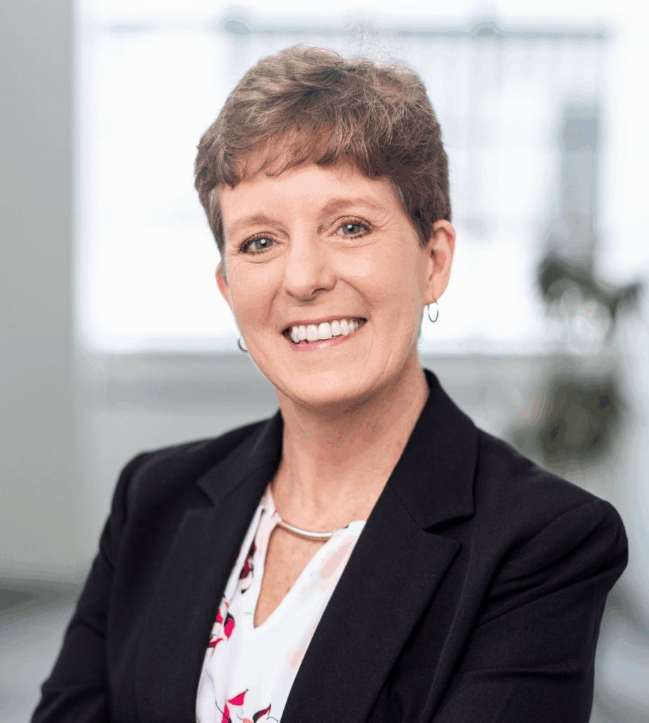 A woman with short brown hair, wearing a black blazer over a white blouse with a floral pattern, smiles at the camera in a bright office setting.