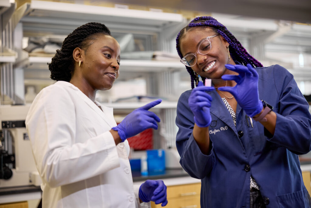 Two scientists wearing lab coats and purple gloves smile and examine a small object together in a laboratory setting, with shelves and scientific equipment in the background.