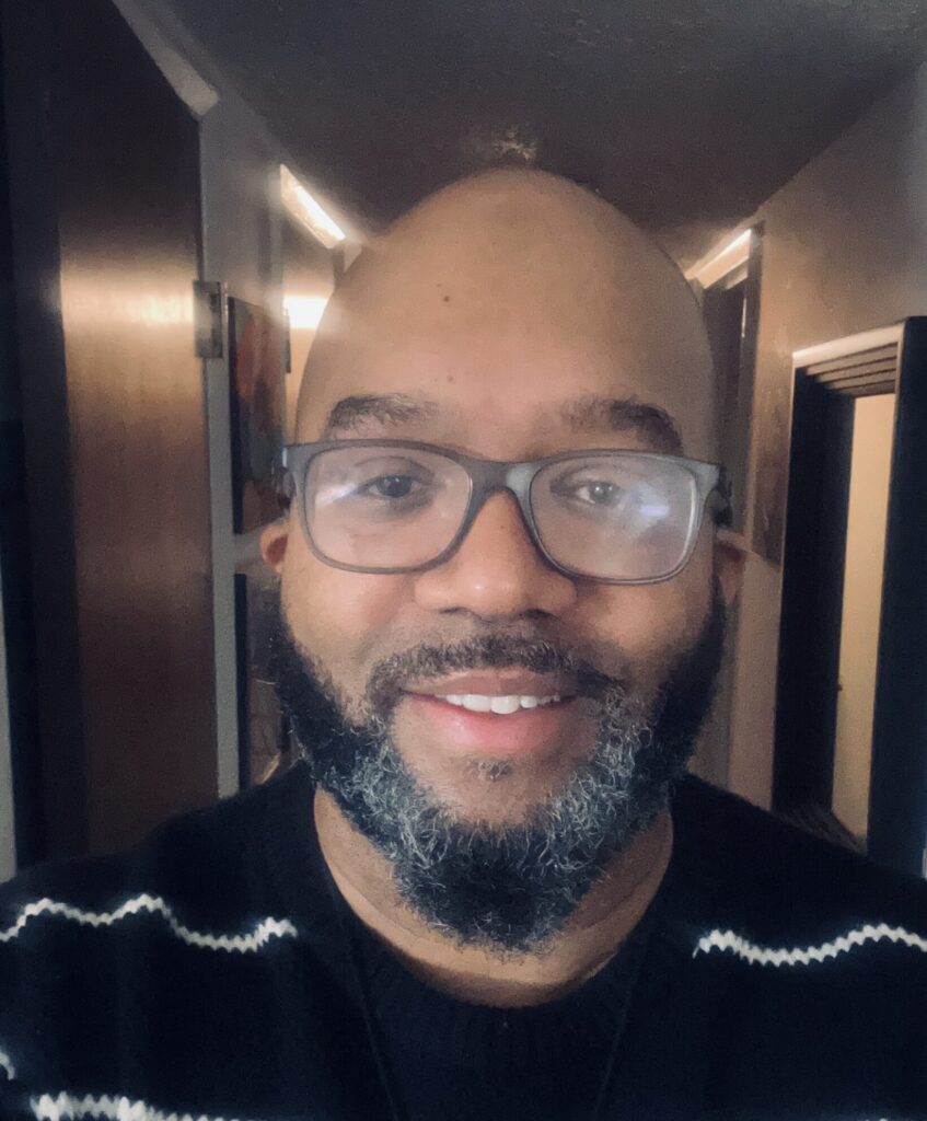 A man with glasses and a salt-and-pepper beard smiles at the camera. He is wearing a black sweater with white stripes and is standing in a hallway with doors on either side.