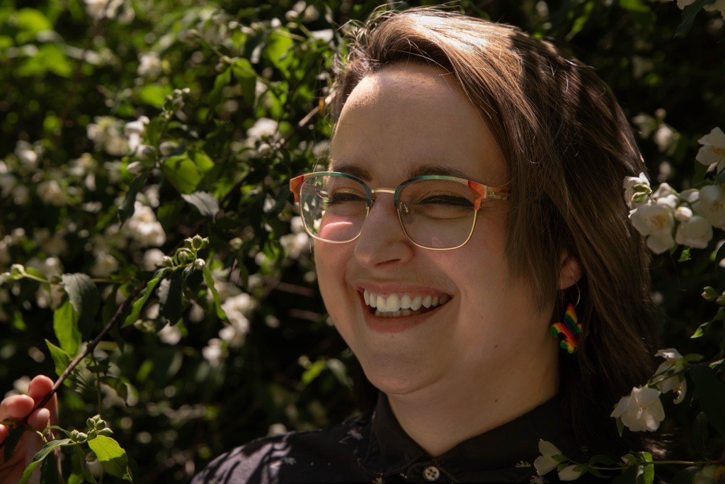 A person with short brown hair, colorful glasses, and rainbow earrings smiles brightly while standing among blooming white flowers and green leaves in sunlight.