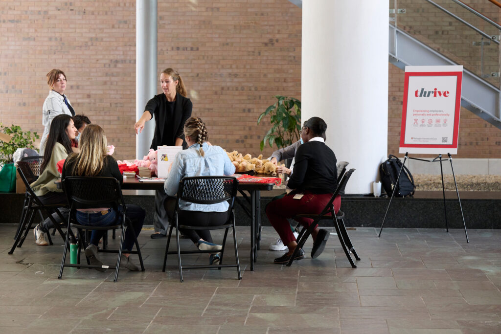 A group of people sit around a table at an indoor event. One woman stands, reaching into a bag. A sign next to the table reads thrive. The setting appears to be a public or community space with a brick wall and pillars.