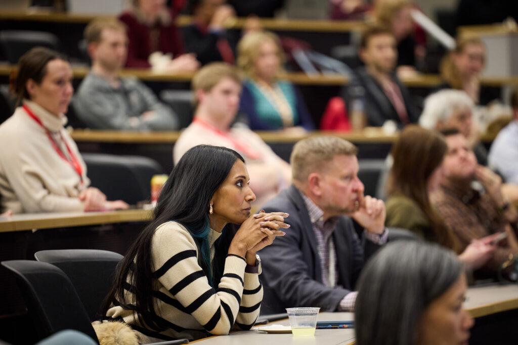 A group of people sit in a lecture hall, attentively listening to a speaker. The focus is on a woman in a striped sweater sitting in the front row, resting her chin on her hands, with others seated behind her.