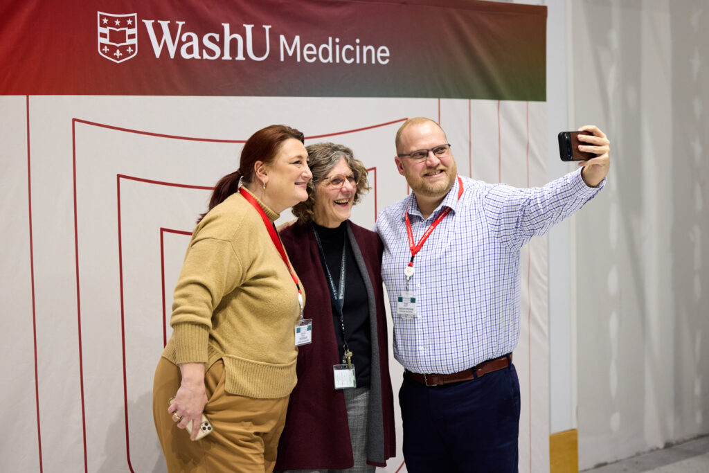 Three people wearing work badges and red lanyards smile and pose for a selfie in front of a WashU Medicine banner. The background suggests an indoor event or gathering.