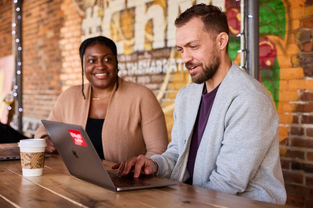 Two people sit at a wooden table with a laptop and coffee cup, smiling and working together in a casual, colorful, brick-walled setting decorated with string lights.