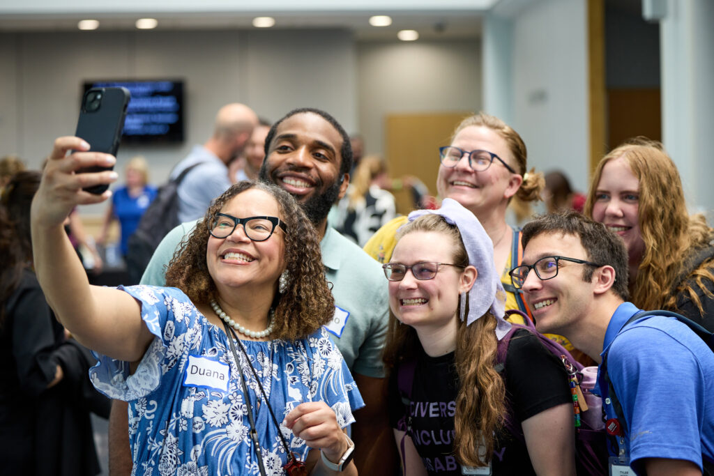 A diverse group of six people, smiling and posing for a selfie at an indoor event. One woman in front holds the phone, and everyone looks happy. They wear nametags and casual clothes; other attendees are visible in the background.