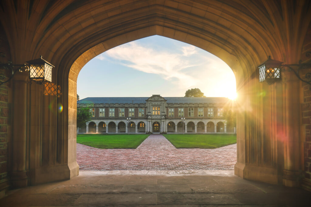 View through an arched stone doorway looking out onto a landscaped courtyard with a historic academic building in the distance, illuminated by the setting sun.